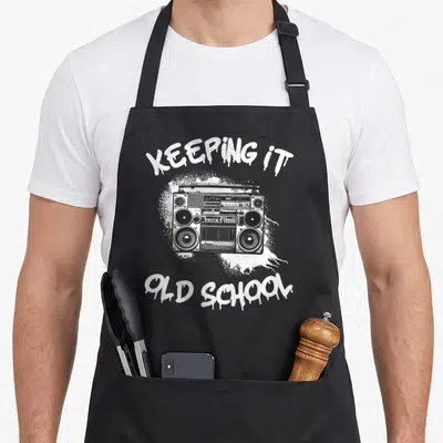 Keeping hip aprons - Black boombox illustration with arched KEEPING IT OLD SCHOOL graffiti text on white splatter — shown on a black cotton canvas apron against a white background