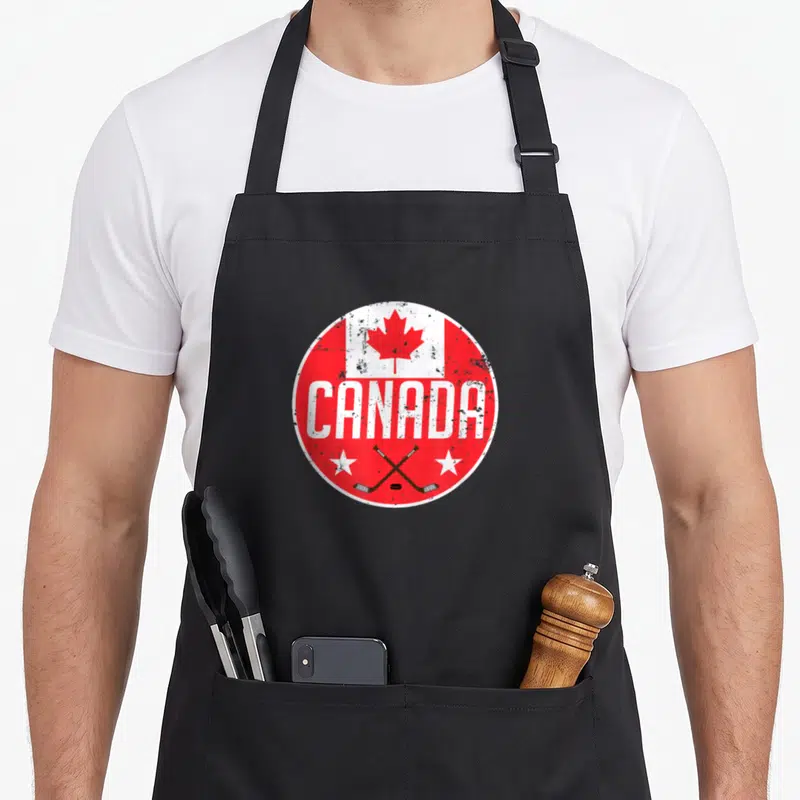 Ice canada aprons - Red white circular logo with CANADA text crossed hockey sticks puck and stars — shown on a black cotton canvas apron against a white background