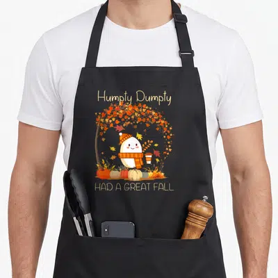 Dumpty cute fall aprons - Cartoon egg character with scarf under autumn tree and pumpkins with text Humpty Dumpty Had — shown on a black cotton canvas apron against a white background