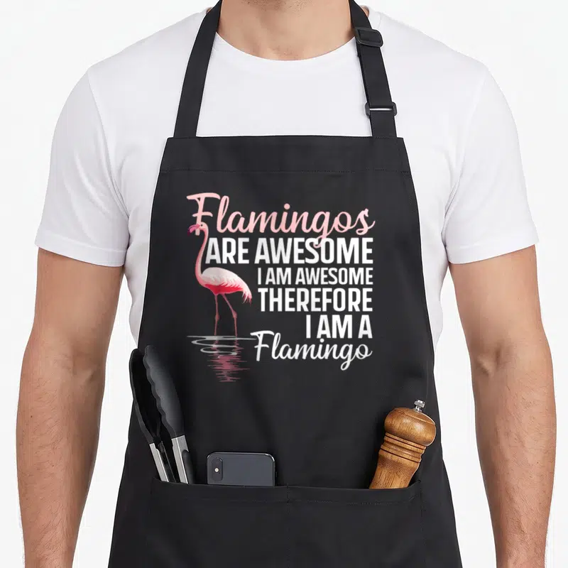 Flock pink flamingo apron - Pink flamingo standing in water next to stacked white and pink text reading Flamingos Are — shown on a black cotton canvas apron against a white background