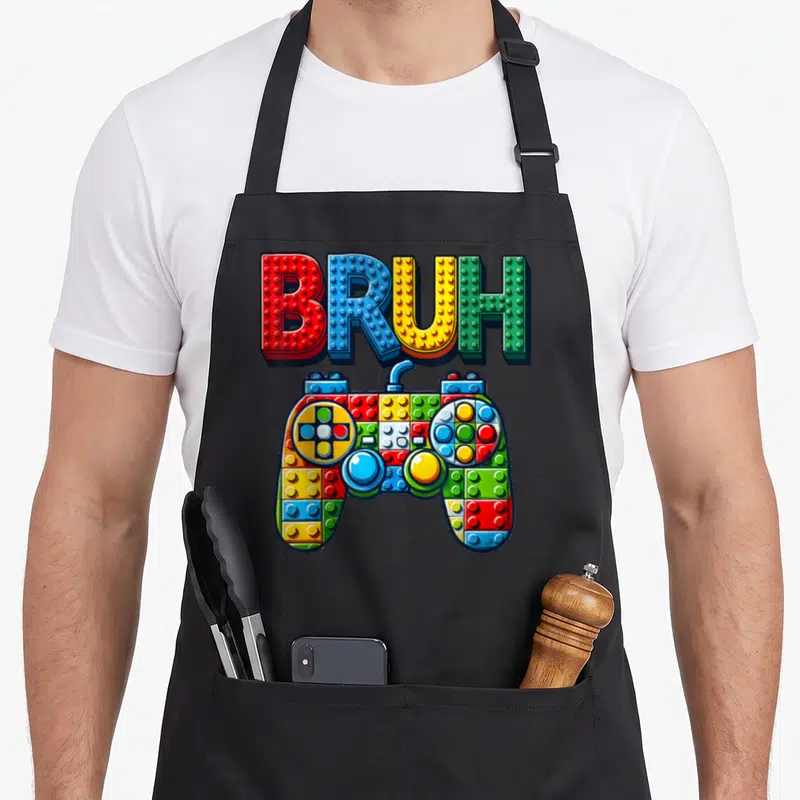 Play building apron - BRUH text and game controller formed from colorful plastic building blocks — shown on a black cotton canvas apron against a white background