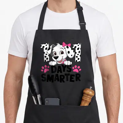 Dalmatian smarter dog apron - Cartoon dalmatian puppy peeking over 101 DAYS SMARTER text flanked by two pink paw prints — shown on a black cotton canvas apron against a white background