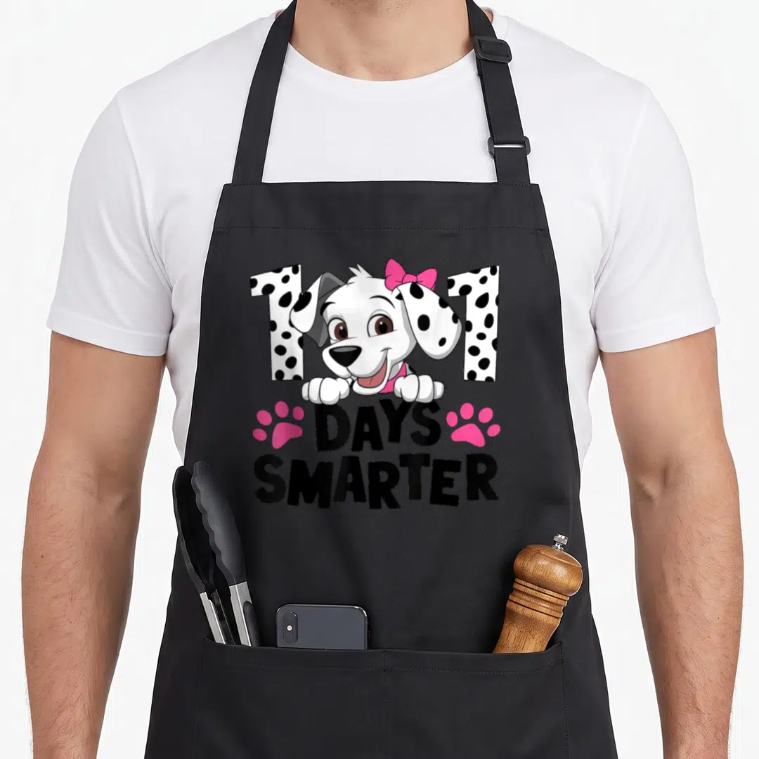 Dalmatian smarter dog apron - Cartoon dalmatian puppy peeking over 101 DAYS SMARTER text flanked by two pink paw prints — shown on a black cotton canvas apron against a white background