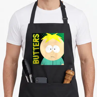 Funny butter apron - Butters Stotch face with vertical yellow text and scattered green dots on black background — shown on a black cotton canvas apron against a white background