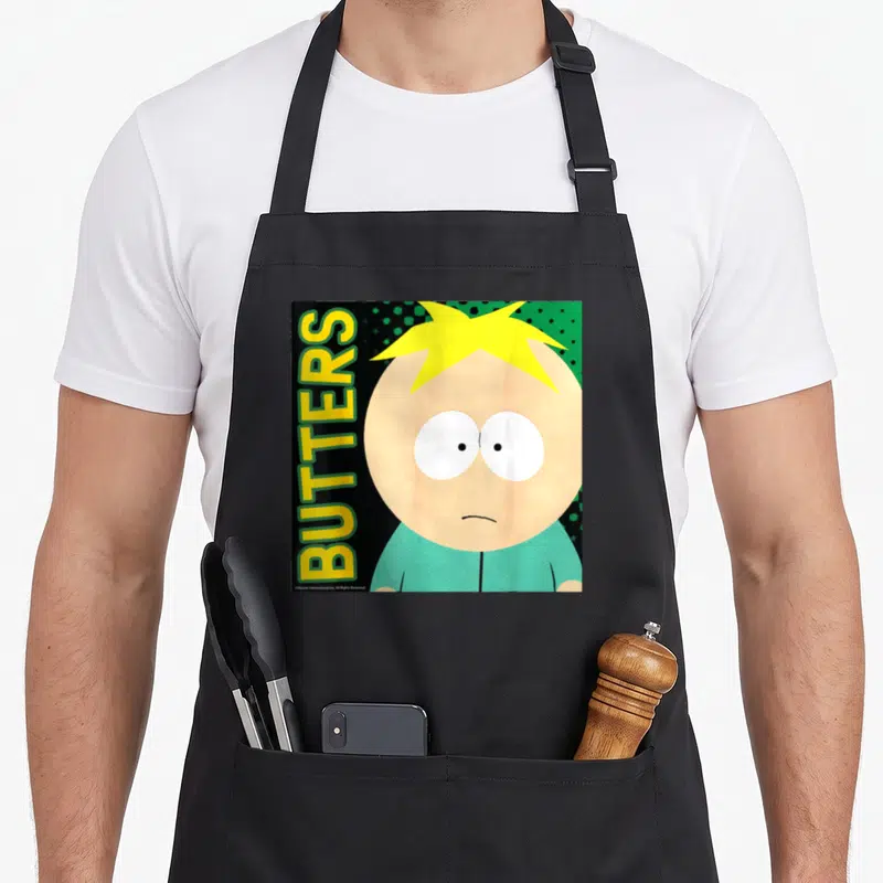 Funny butter apron - Butters Stotch face with vertical yellow text and scattered green dots on black background — shown on a black cotton canvas apron against a white background