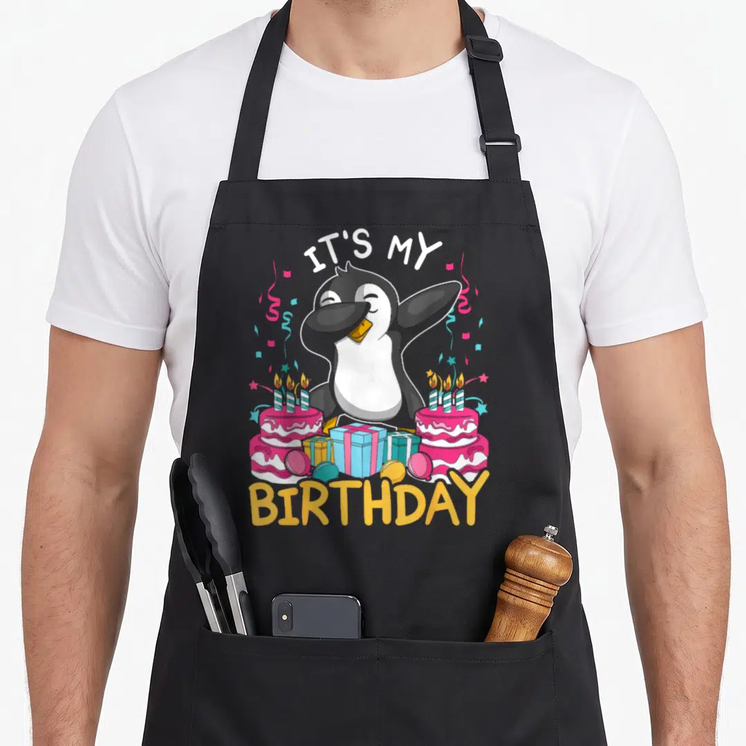 Animal cake apron - Dabbing penguin between two pink birthday cakes with IT'S MY BIRTHDAY text and confetti — shown on a black cotton canvas apron against a white background