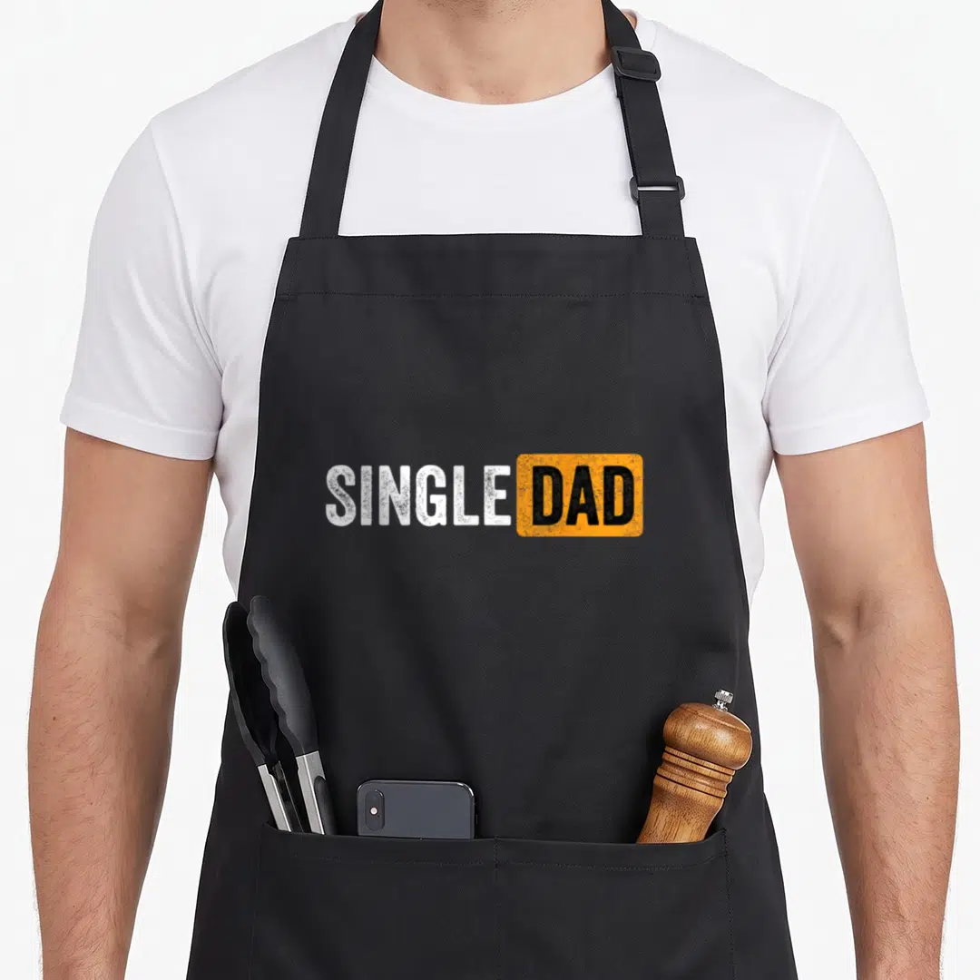 Parent single apron - SINGLE DAD text in white and black inside orange rectangle with distressed finish — shown on a black cotton canvas apron against a white background