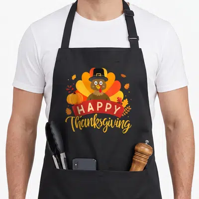 Feast thanksgiving apron - Cartoon turkey in hat with red HAPPY banner, pumpkin, and autumn leaves — shown on a black cotton canvas apron against a white background
