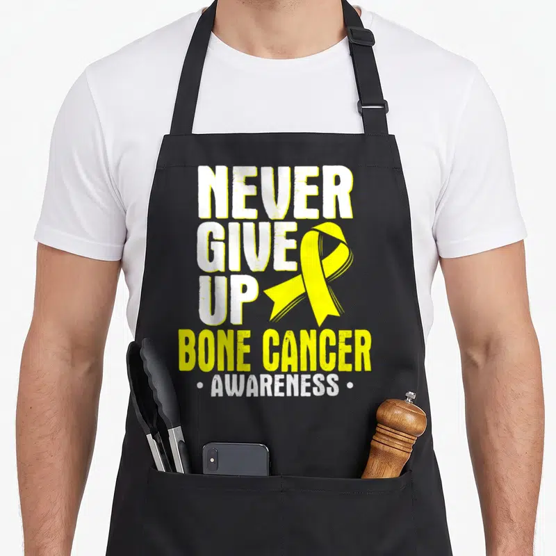 Ewing bone cancer awareness apron - Yellow ribbon overlapping white NEVER GIVE UP text with BONE CANCER AWARENESS below — shown on a black cotton canvas apron against a white background