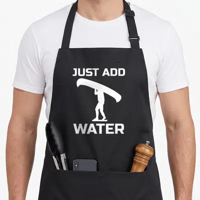Canoeing canoe guy apron - White silhouette of person carrying surfboard between JUST ADD and WATER text — shown on a black cotton canvas apron against a white background