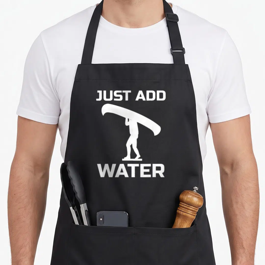 Canoeing canoe guy apron - White silhouette of person carrying surfboard between JUST ADD and WATER text — shown on a black cotton canvas apron against a white background