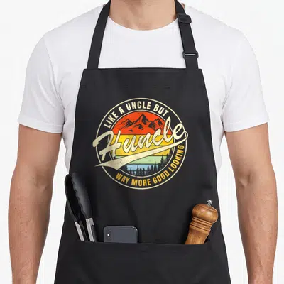 Uncle modern apron - Retro circular Huncle badge with sunset mountains and trees in red orange yellow blue — shown on a black cotton canvas apron against a white background