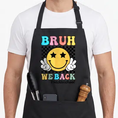 Funny bruh teacher apron - Yellow smiley face with star eyes flanked by peace hands under BRUH WE BACK text — shown on a black cotton canvas apron against a white background