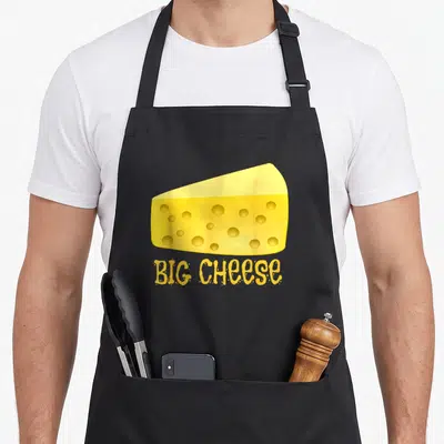 Cheese boss apron - Yellow swiss cheese wedge above bold big cheese text in cartoon style — shown on a black cotton canvas apron against a white background
