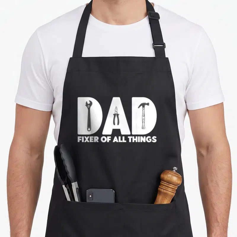 Tool woodworking apron - White DAD letters contain wrench pliers hammer. Below reads Fixer of All Things — shown on a black cotton canvas apron against a white background