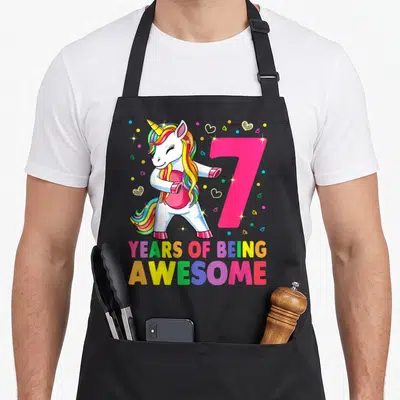 Flossing 7th birthday apron - Dancing unicorn beside pink number 7 with rainbow text and scattered hearts — shown on a black cotton canvas apron against a white background