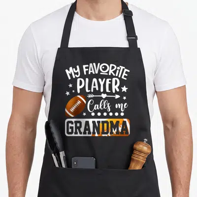 Football player grandma aprons - Stacked white text My Favorite Player Calls Me Grandma with brown footballs and stars — shown on a black cotton canvas apron against a white background