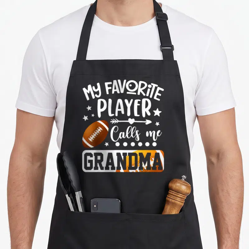 Football player grandma aprons - Stacked white text My Favorite Player Calls Me Grandma with brown footballs and stars — shown on a black cotton canvas apron against a white background