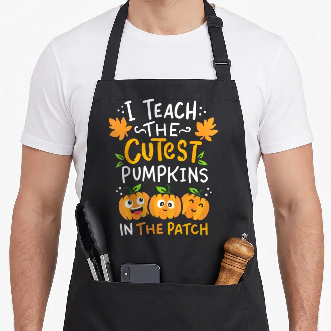 Halloween kindergarten teacher apron - I Teach the Cutest Pumpkins in the Patch text with three smiling cartoon — shown on a black cotton canvas apron against a white background