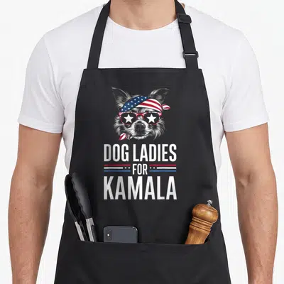 Kamala dog apron - Cartoon grey dog with patriotic bandana and star sunglasses above DOG LADIES FOR KAMALA text — shown on a black cotton canvas apron against a white background