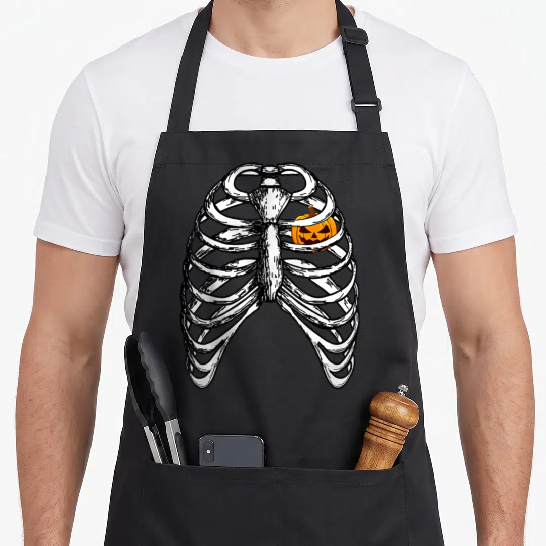 Pumpkin rib halloween apron - White ribcage line art with orange jack-o-lantern inside chest cavity — shown on a black cotton canvas apron against a white background
