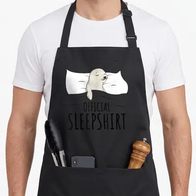 Sleepshirt lion apron - Cartoon seal sleeping on white pillow under grey blanket with official sleepshirt text below — shown on a black cotton canvas apron against a white background