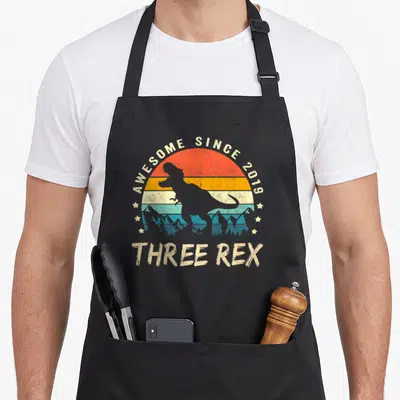 Three t rex apron - Retro striped sun with T-Rex silhouette and AWESOME SINCE 2019 text — shown on a black cotton canvas apron against a white background
