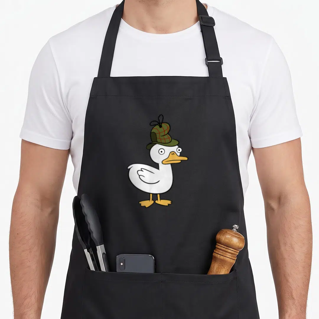Channel duck apron - White cartoon duck wearing a green and red plaid hat with orange beak and feet — shown on a black cotton canvas apron against a white background