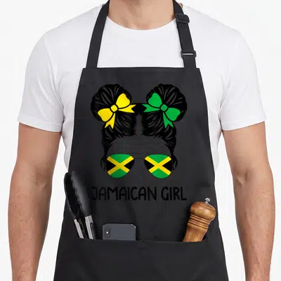 Jamaican messy aprons - Cartoon girl head with buns, yellow and green bows, flag sunglasses, JAMAICAN GIRL text below — shown on a black cotton canvas apron against a white background