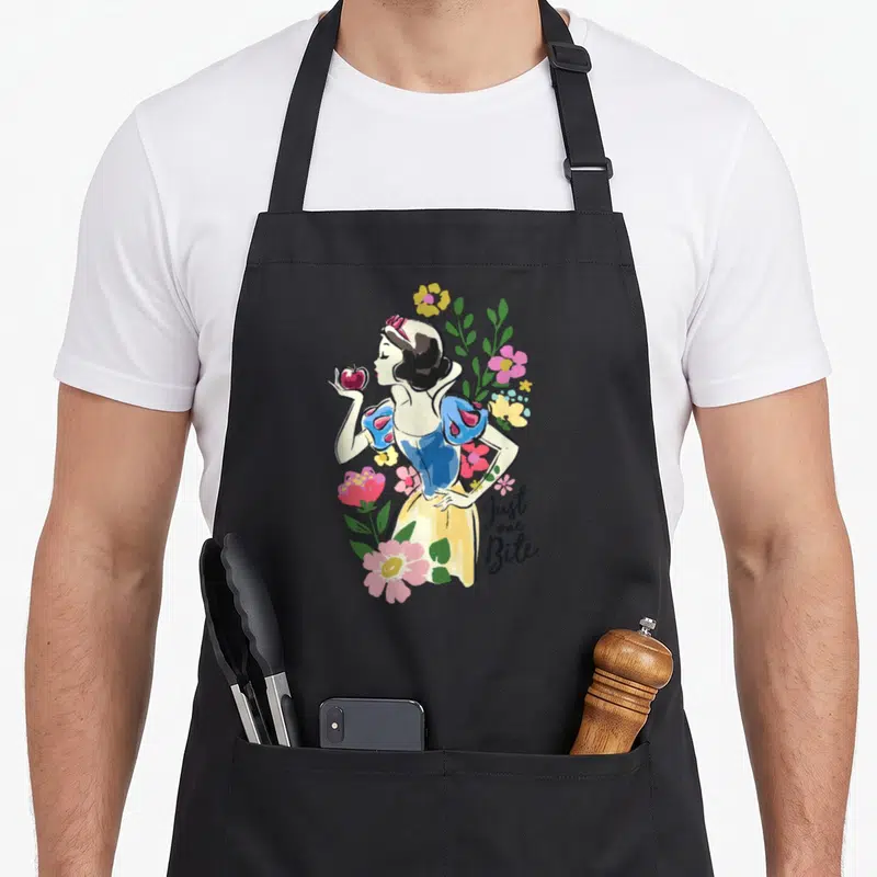 Bite floral apron - Snow White holding red apple surrounded by pink yellow green flowers with stacked text — shown on a black cotton canvas apron against a white background