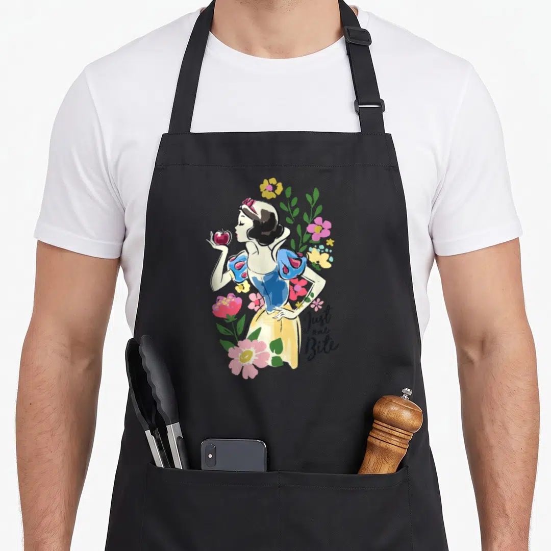 Bite floral apron - Snow White holding red apple surrounded by pink yellow green flowers with stacked text — shown on a black cotton canvas apron against a white background