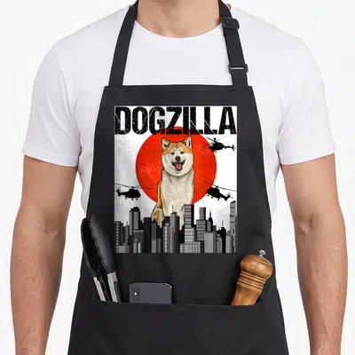 Japanese akita apron - DOGZILLA text over Shiba Inu, red sun, black helicopters, and city skyline in cartoon style — shown on a black cotton canvas apron against a white background