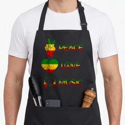 Peace pride apron - PEACE LOVE MUSIC text with rasta striped peace hand heart and headphones in distressed style — shown on a black cotton canvas apron against a white background