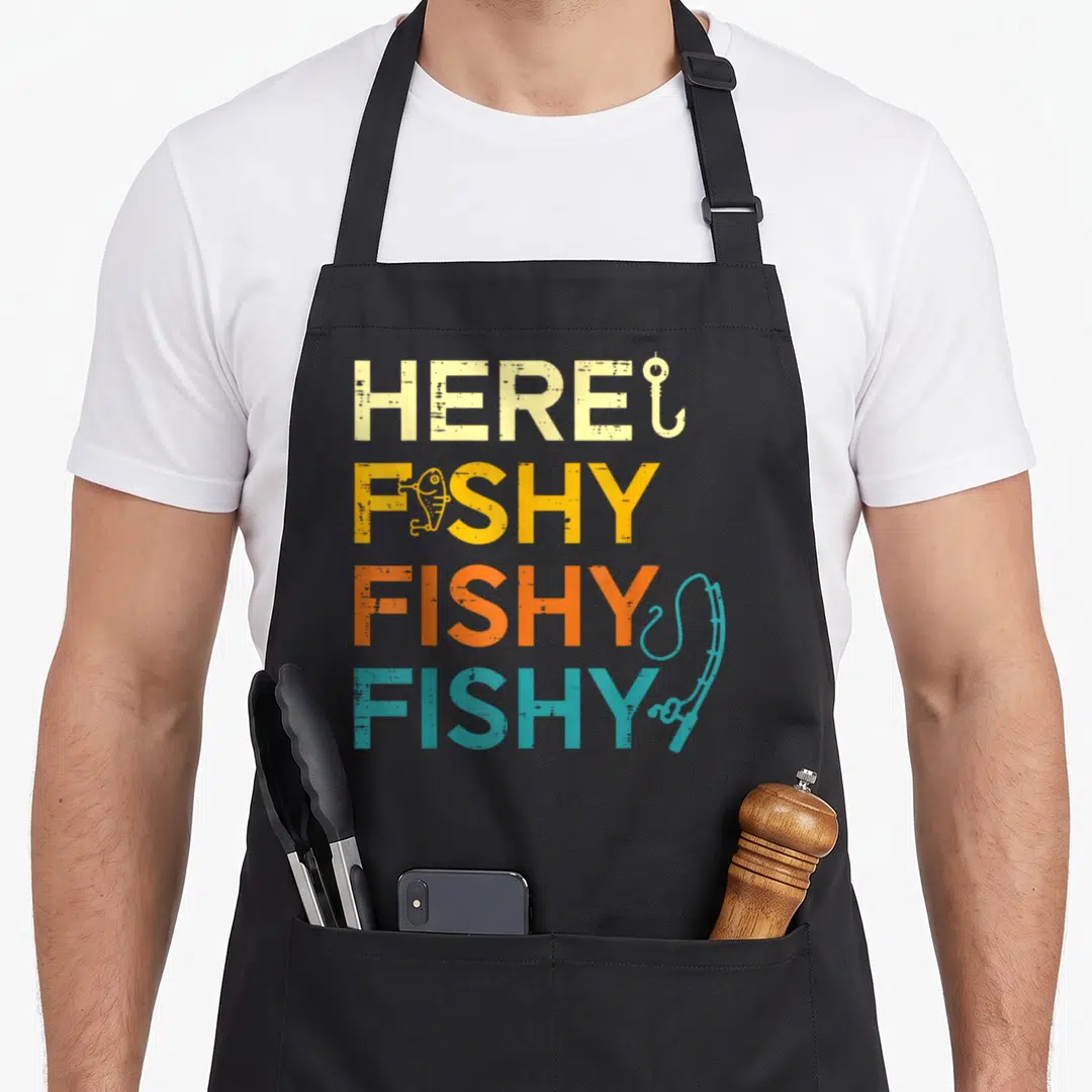 Fishy fisherman apron - Stacked text HERE FISHY FISHY FISHY in beige yellow orange teal with hook lure and rod — shown on a black cotton canvas apron against a white background