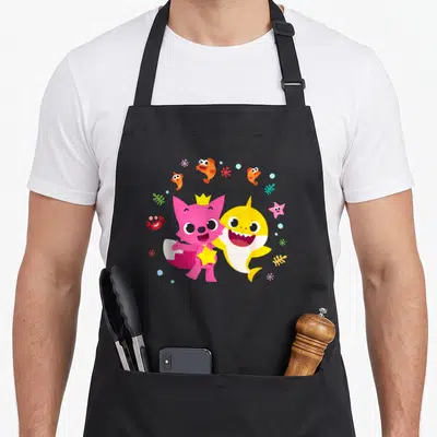 Shark baby apron - Yellow and pink cartoon sharks standing with orange fish, red crab, and blue bubbles — shown on a black cotton canvas apron against a white background