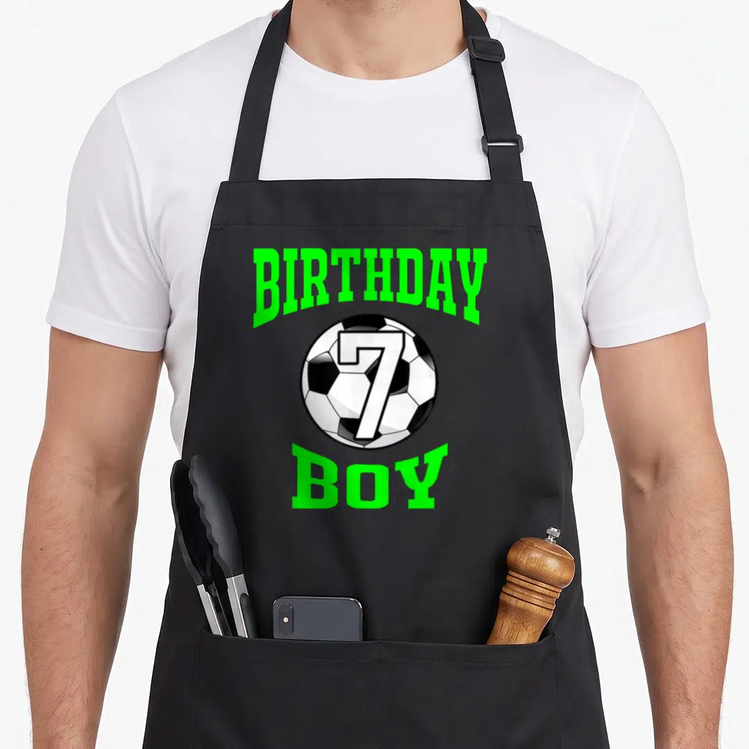 7th soccer birthday apron - Cartoon soccer ball with number 7 and arched BIRTHDAY BOY text in lime green — shown on a black cotton canvas apron against a white background