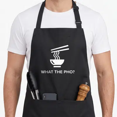 Funny vietnamese pho apron - White graphic of steaming pho bowl with chopsticks and text What The Pho? — shown on a black cotton canvas apron against a white background