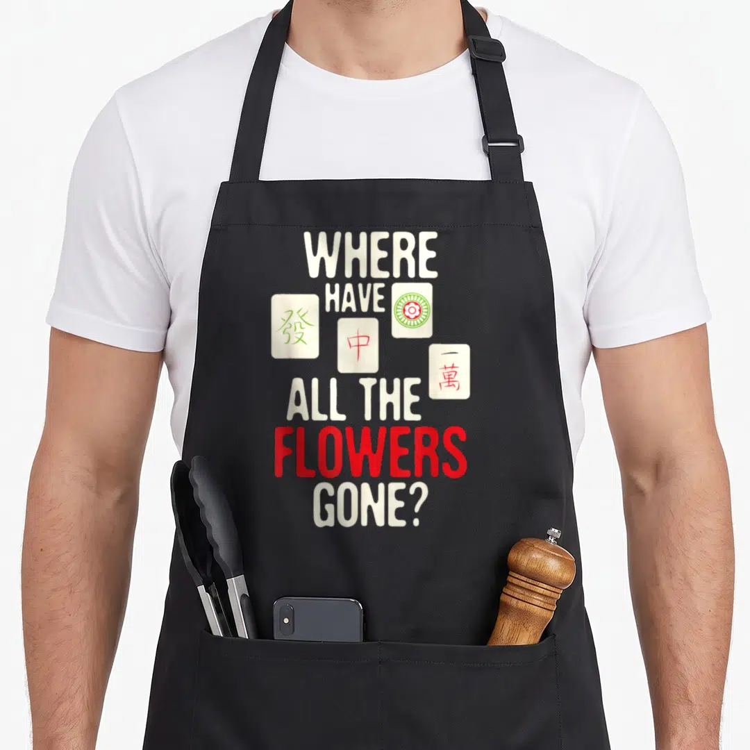 Flower mahjong apron - Stacked text WHERE HAVE ALL THE FLOWERS GONE? with four mahjong tiles in red and green markings — shown on a black cotton canvas apron against a white background