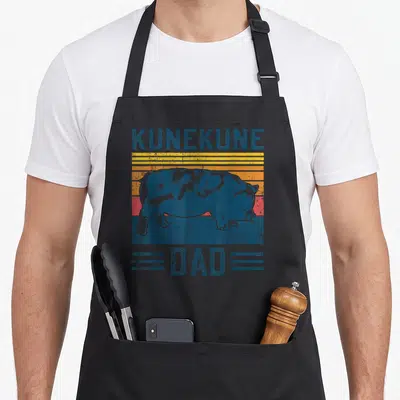 Breed farming apron - Retro blue pig silhouette on yellow orange red stripes with KUNEKUNE DAD text — shown on a black cotton canvas apron against a white background