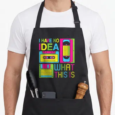 Cute 80s chef outfit with apron - Pink yellow blue and green cassette tape with text I Have No Idea What This Is — shown on a black cotton canvas apron against a white background