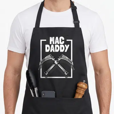 Laryngoscope mac apron - MAC DADDY text above two crossed laryngoscopes in white grey black cartoon style — shown on a black cotton canvas apron against a white background