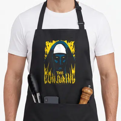 Conjuring horror movie apron - Distressed nun face with blue hood and yellow flames, stacked text below — shown on a black cotton canvas apron against a white background