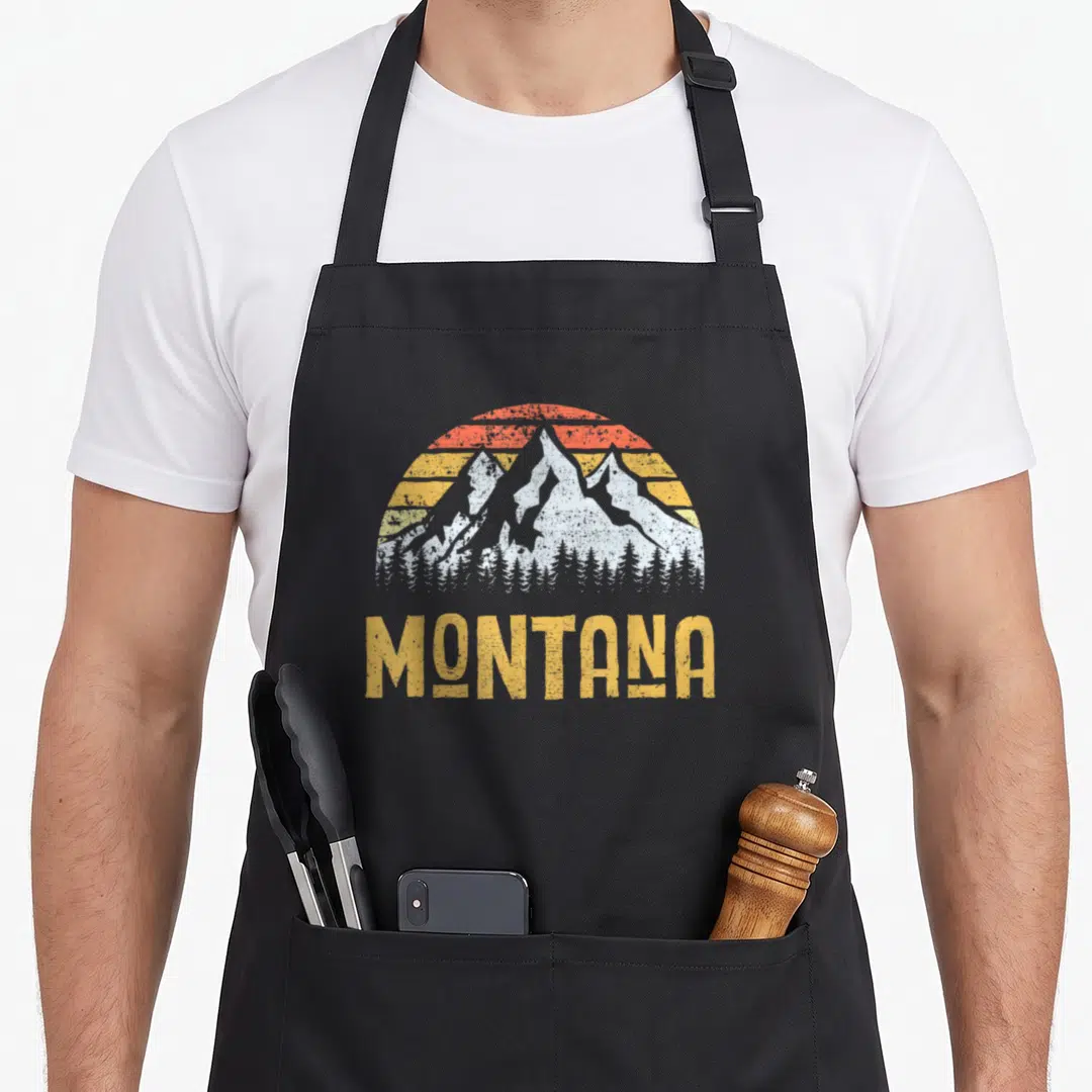 Mount montana apron - Retro white mountain silhouette behind orange and yellow striped sun with MONTANA text below — shown on a black cotton canvas apron against a white background