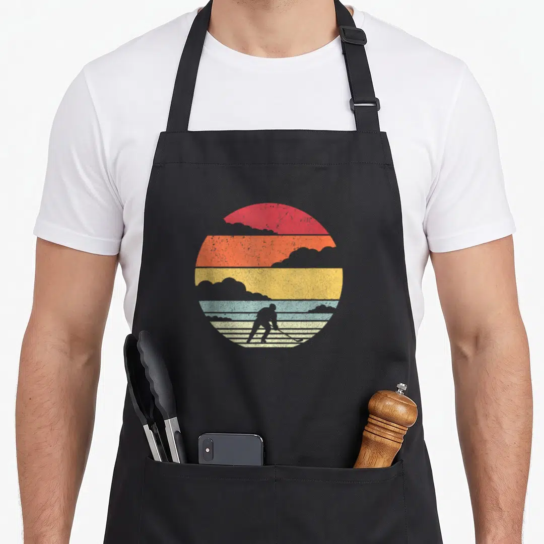 Hockey frappe apron - Retro striped sun with hockey player silhouette in red orange yellow blue grey clouds — shown on a black cotton canvas apron against a white background