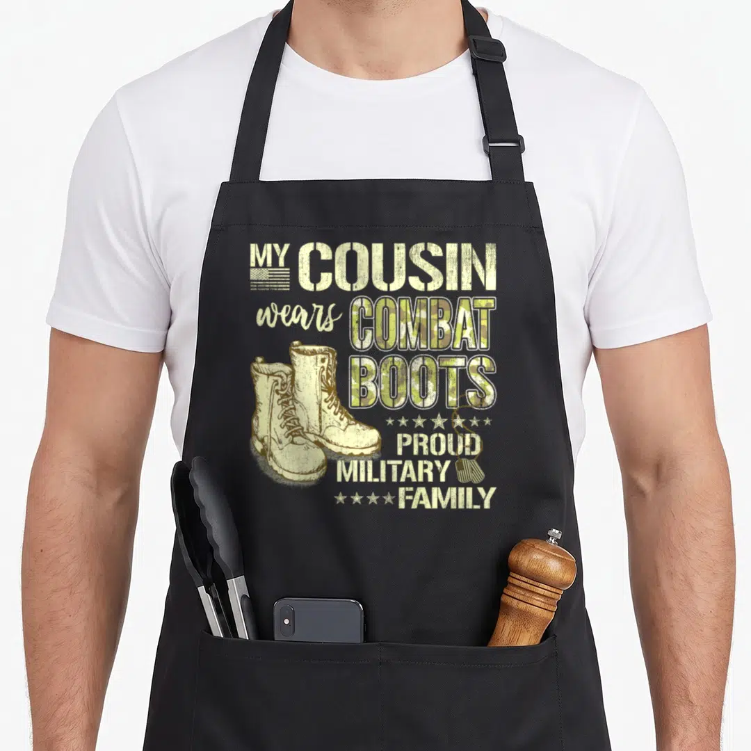Cousin tag apron - My Cousin Wears Combat Boots text with tan boots, olive camo letters, stars, and dog tags — shown on a black cotton canvas apron against a white background