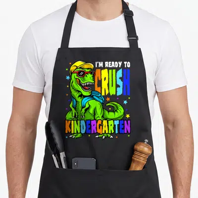 Ready dinosaur apron - Cartoon T-Rex in green with yellow cap red sunglasses blue backpack and multicolored text I'M — shown on a black cotton canvas apron against a white background