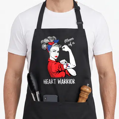 Warrior organ donation apron - Rosie the Riveter flexing arm with red blue ribbon and Mom of a Heart Warrior text — shown on a black cotton canvas apron against a white background