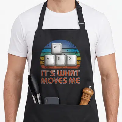 Wasd retro gaming apron - Four white keyboard keys in a T-shape over a pixelated rainbow arc with orange text IT'S WHAT — shown on a black cotton canvas apron against a white background