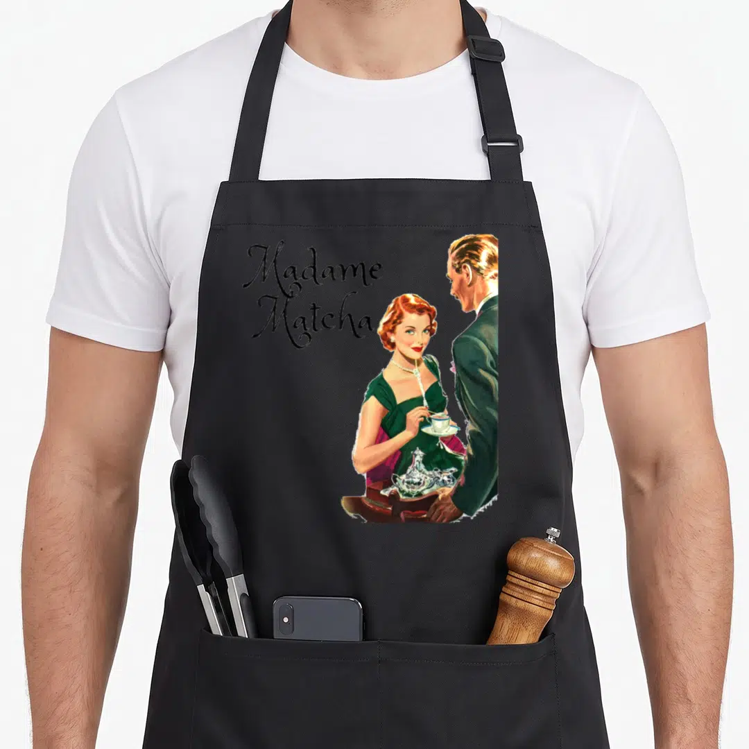 Tea matcha apron - Woman in green dress holds teacup beside man in suit with Madame Matcha text stacked left — shown on a black cotton canvas apron against a white background
