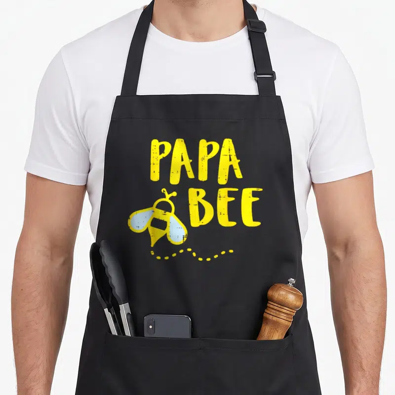 Bee grill daddy apron - Cartoon bee with light blue wings next to yellow PAPA BEE text and dotted flight path — shown on a black cotton canvas apron against a white background